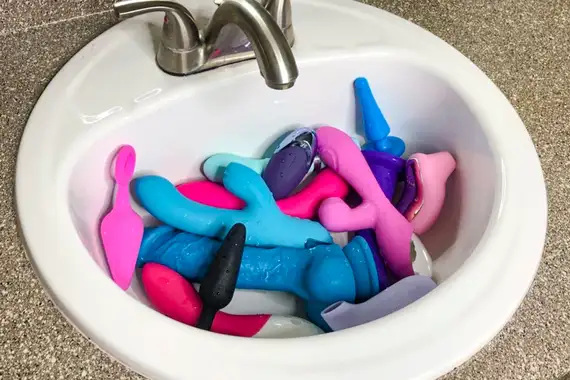 A variety of sex toys being cleaned safely with soap and water in a clean sink.